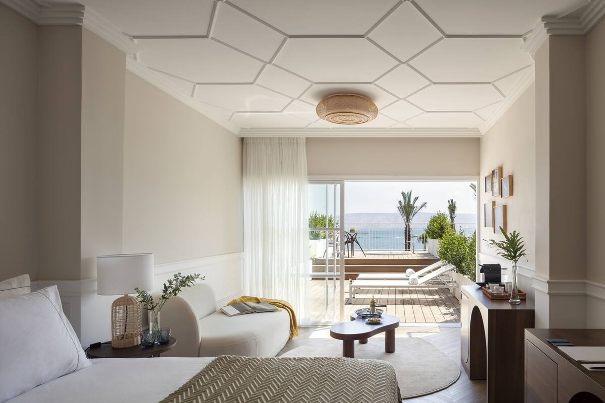 A hotel room with a bed, a desk, a sofa, a balcony with a view of the sea, and a ceiling with a hexagonal pattern.