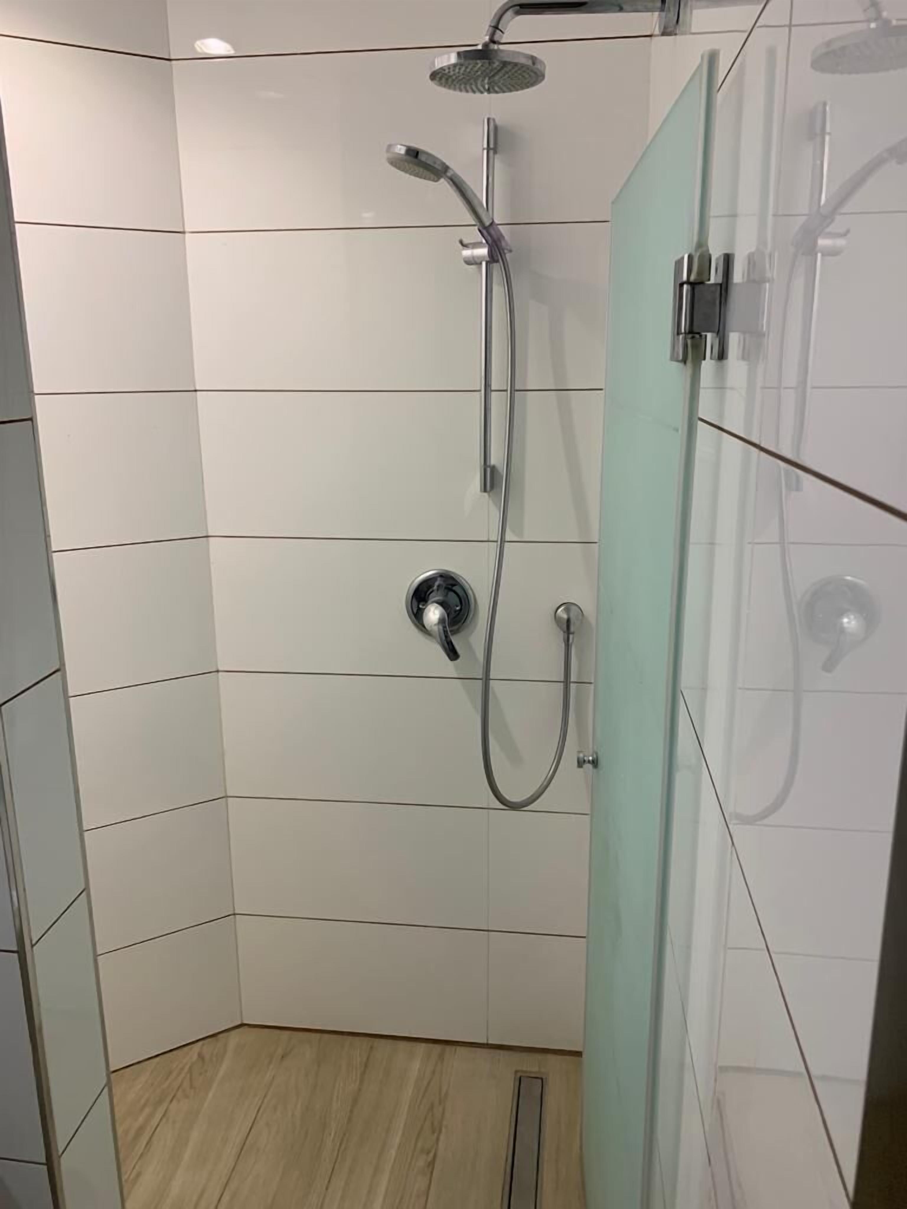 Ducha en esquina con mampara de vidrio, paredes de azulejos blancos y suelo de madera.