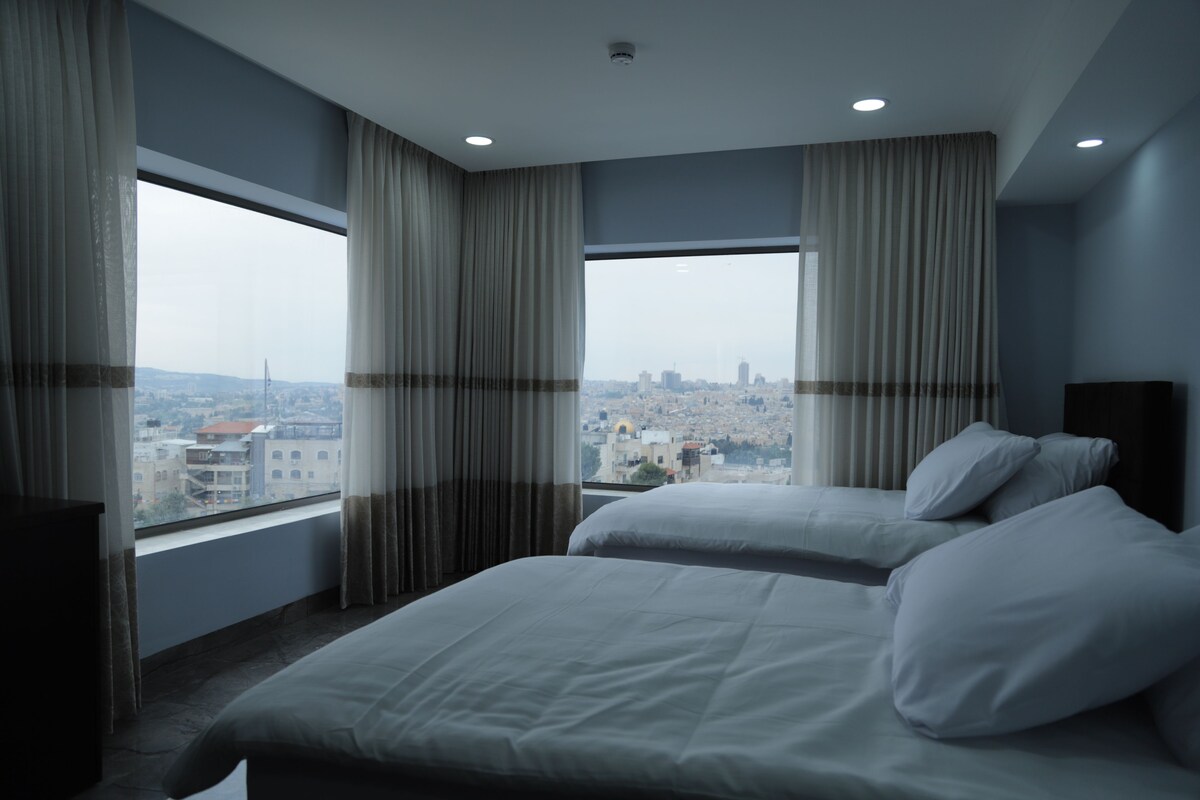A hotel room with two beds, large windows with curtains, a city view, and a flat-screen TV.