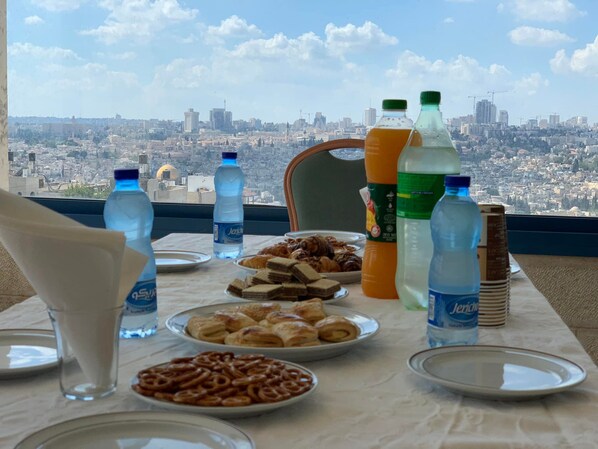 A table with various food items and drinks, a chair, and a view of a cityscape through a window. - Mount Of Olives Hotel (Jerusalem)