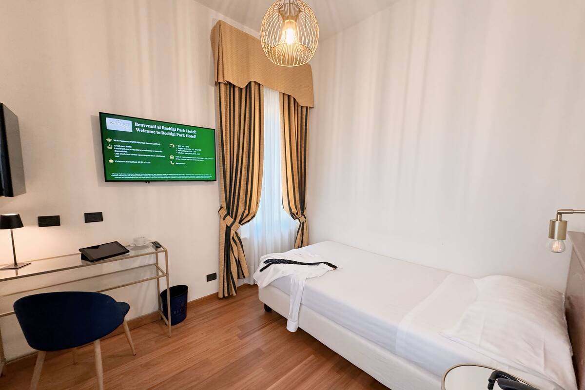 A hotel room with a bed, a desk with a computer, a chair, a television, and a sign with hotel information.