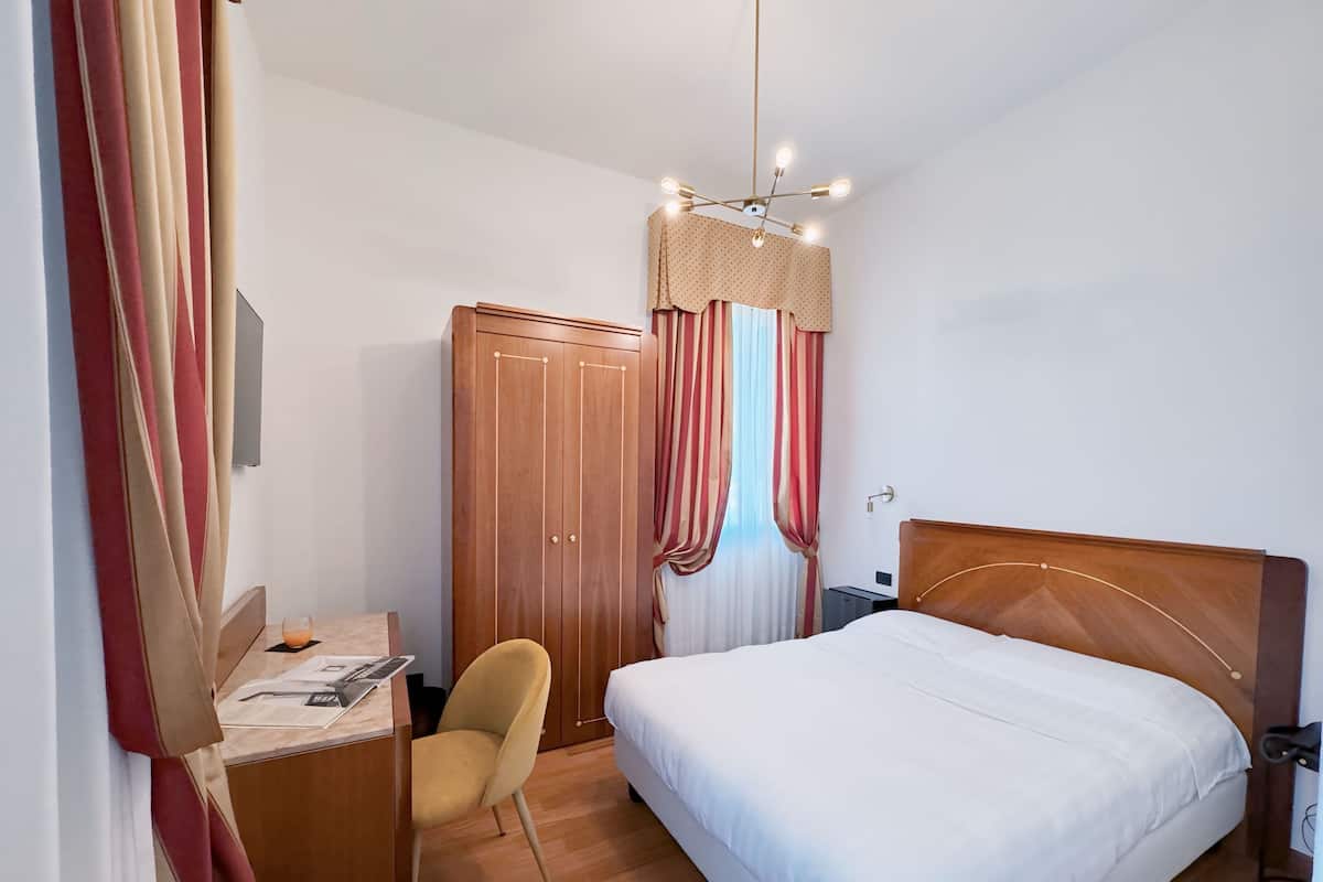 A hotel room with a wooden bed, a desk with a chair, a wardrobe, and a window with curtains.