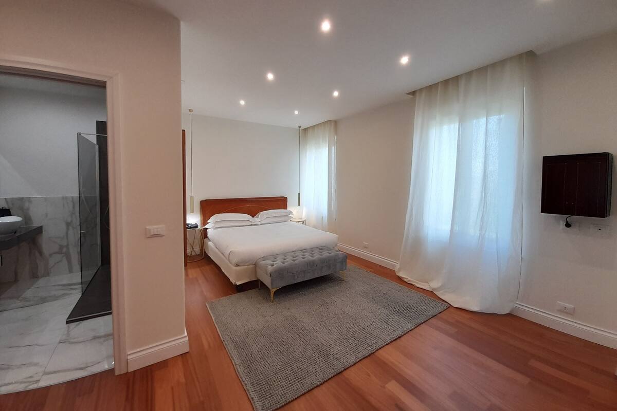 A bedroom with a bed, a bedside table, a TV mounted on the wall, and a bathroom with a glass shower enclosure.