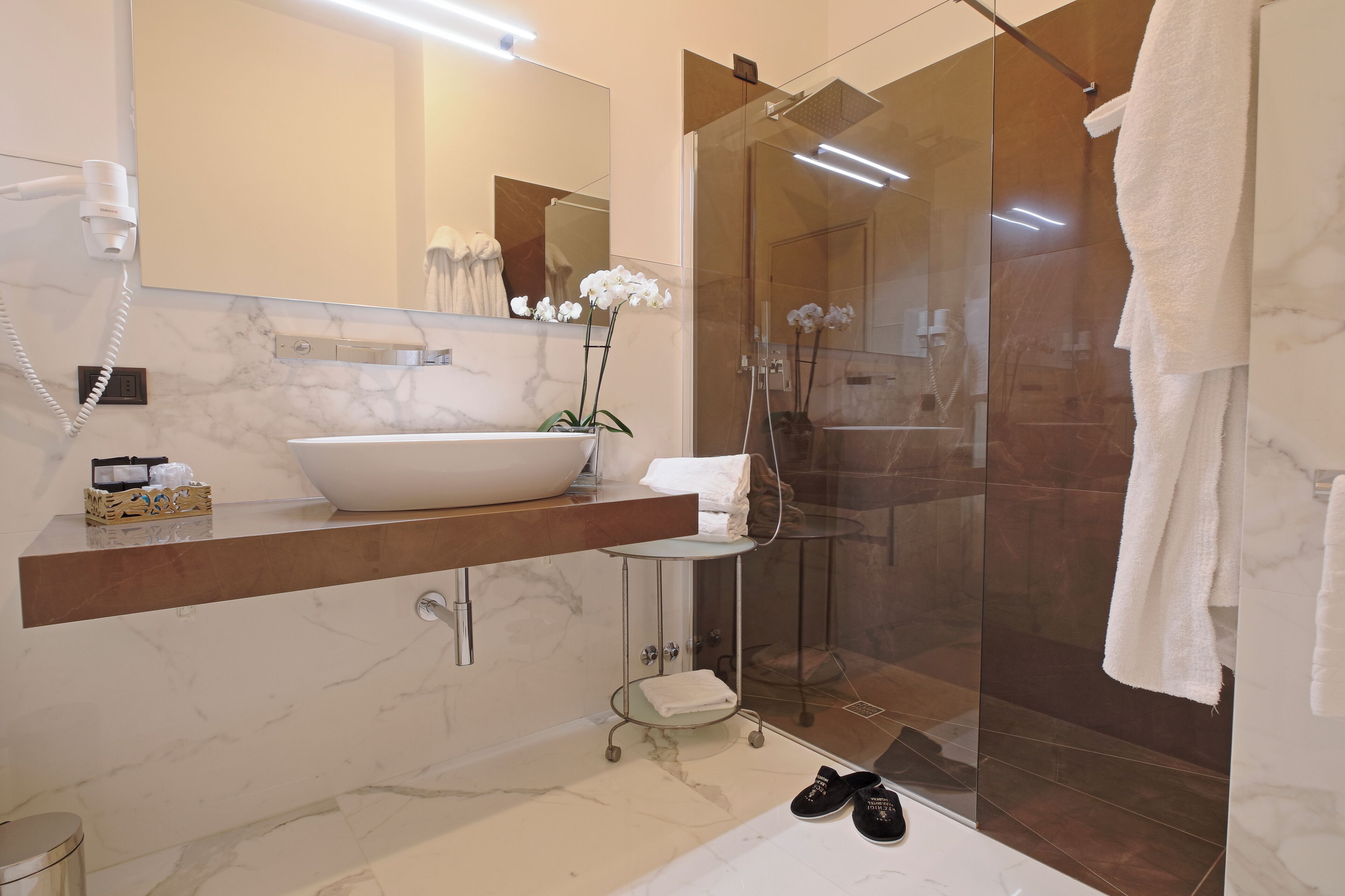 A modern bathroom with a large white sink, a glass shower enclosure, and a marble-tiled floor.
