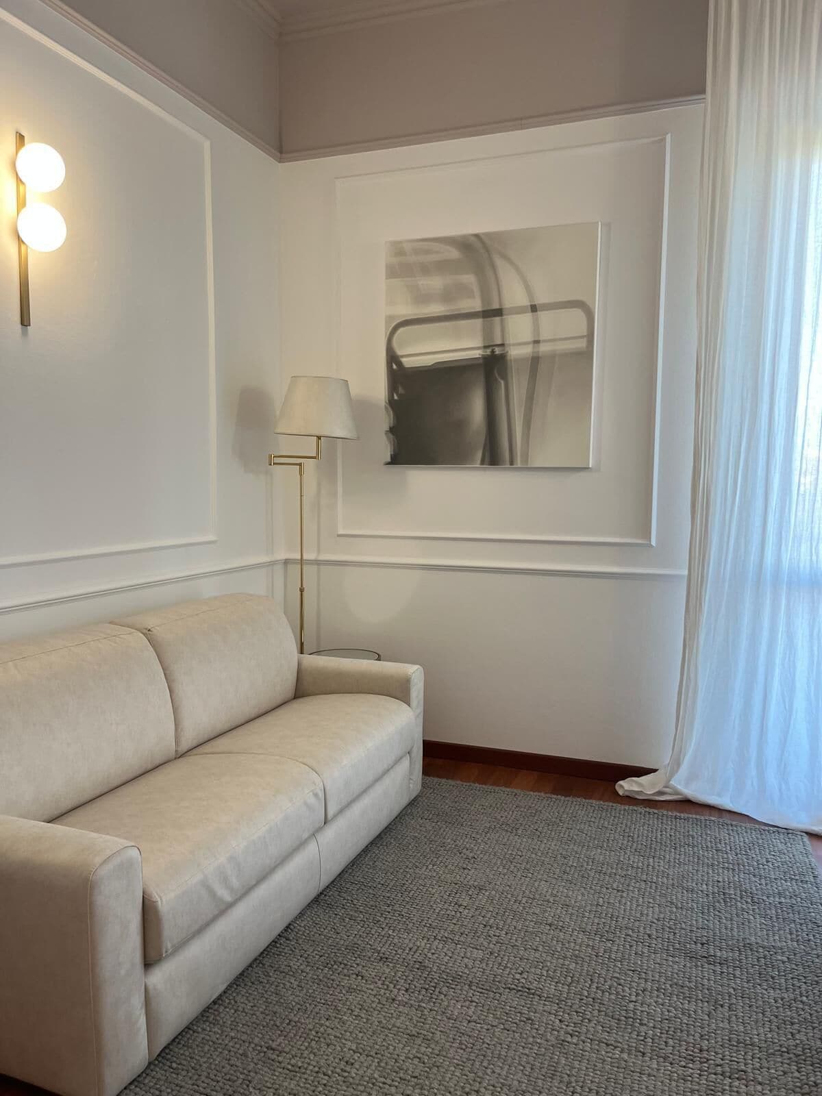 Ein Zimmer mit einem beigen Sofa, einer Stehlampe, einem Gemälde und einem Fenster mit durchsichtigen Vorhängen.