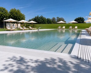 Piscina con sillas de descanso y sombrillas, rodeada por una terraza de madera y follaje.