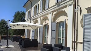 An outdoor seating area with wicker furniture and a large umbrella, set against a building with arched windows and shutters. - Rechigi Park Hotel (Modena)