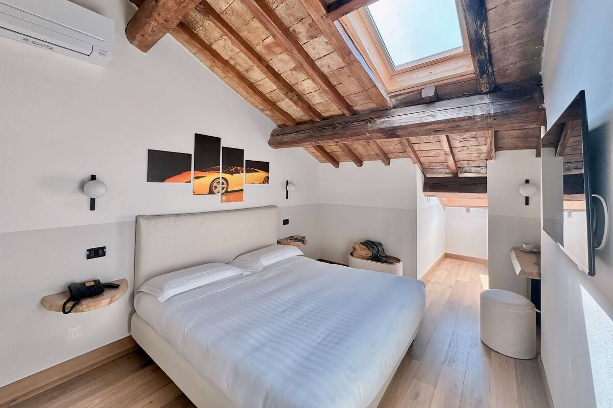 A bedroom with a large bed, wooden beams, a skylight, a wall-mounted artwork, bedside tables, and a small round stool.