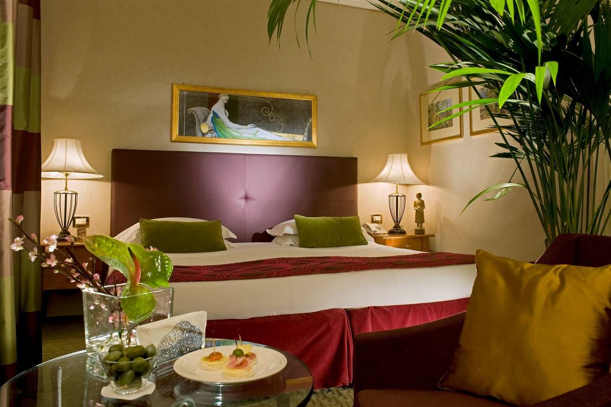 A hotel room with a large bed, two bedside lamps, a nightstand with a lamp, a framed picture on the wall, and a potted plant.