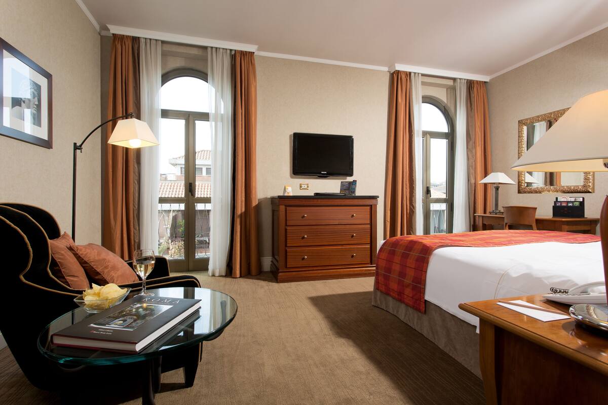 A hotel room with a bed, a television, a desk with a lamp, a chair, and a window with curtains.