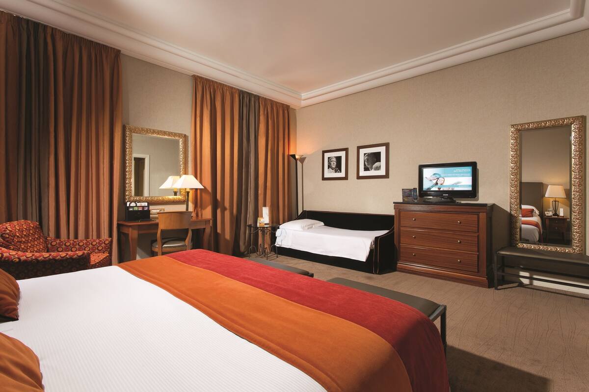 A hotel room with a large bed, a desk with a lamp, a television, and two framed pictures on the wall.
