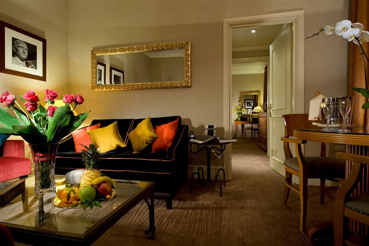 A living room with a black sofa, colorful cushions, a fruit tray, a vase with flowers, a mirror, and a dining area with a table and chairs.