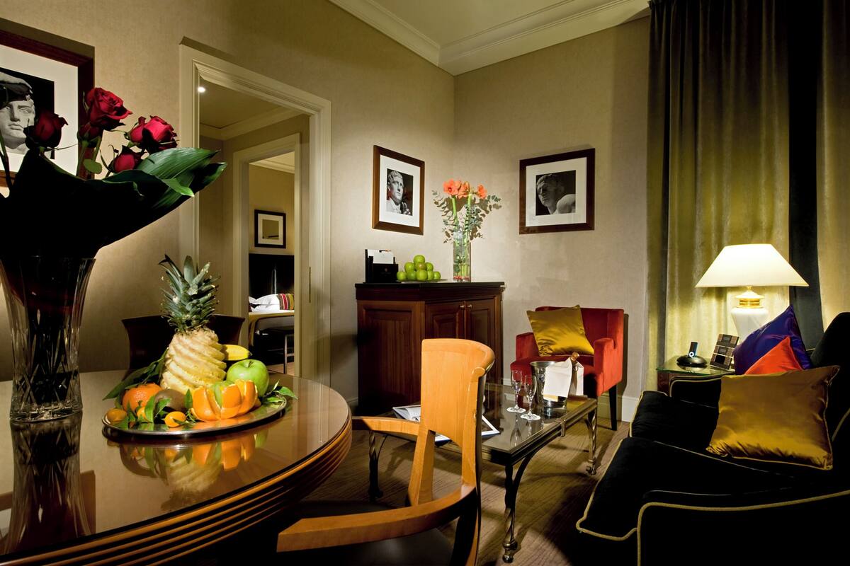 A well-lit living room with a dining area, featuring a round table with fruits, a vase with flowers, and a sofa with cushions.