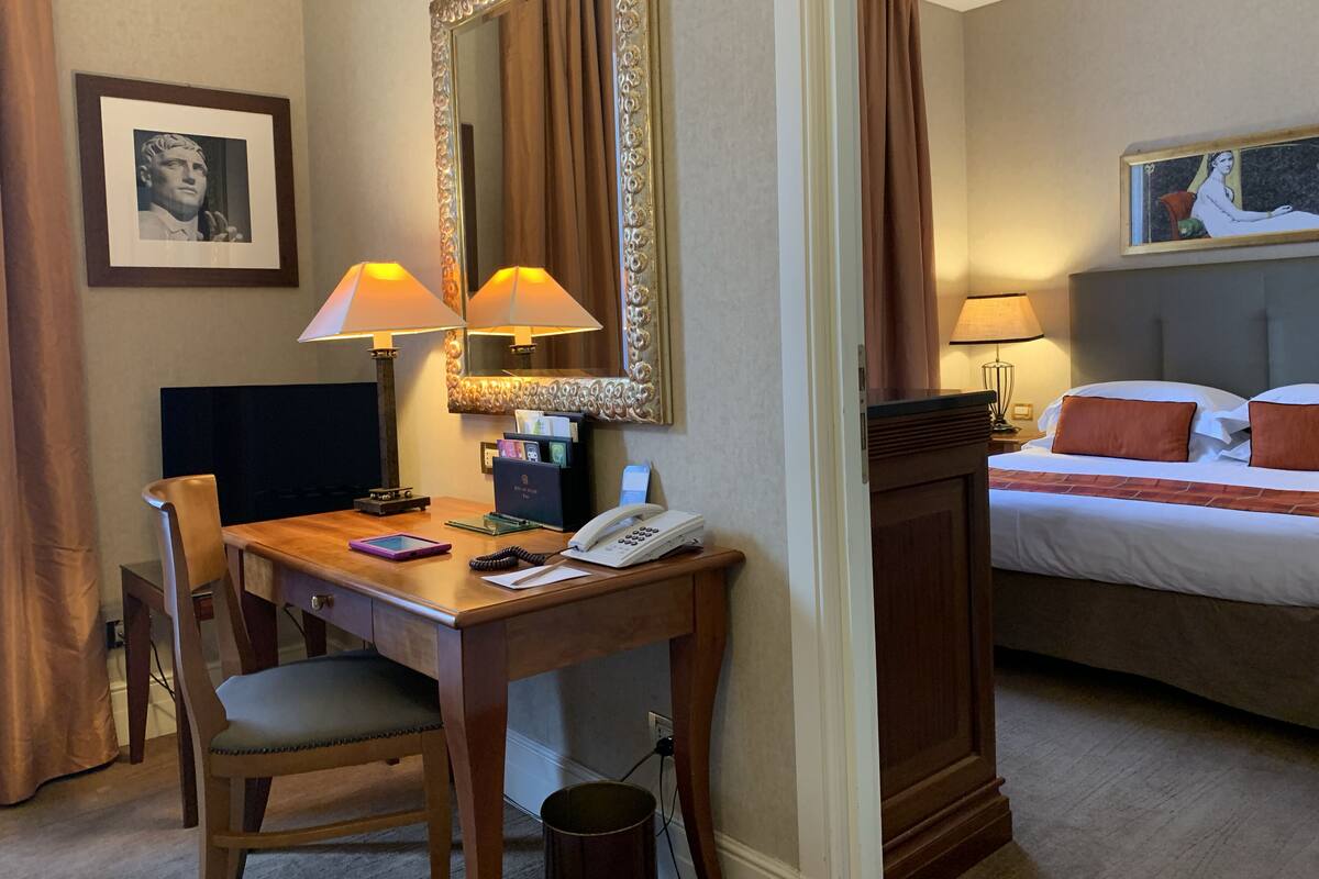 A hotel room with a desk, chair, two lamps, a bed with pillows, a framed picture on the wall, and a mirror.
