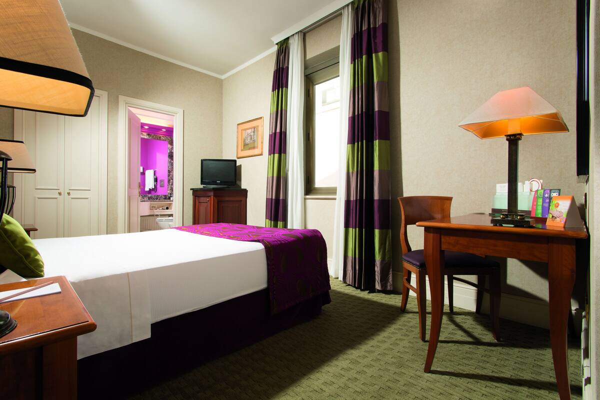 A hotel room with a bed, a desk, a chair, a television, and a lamp.