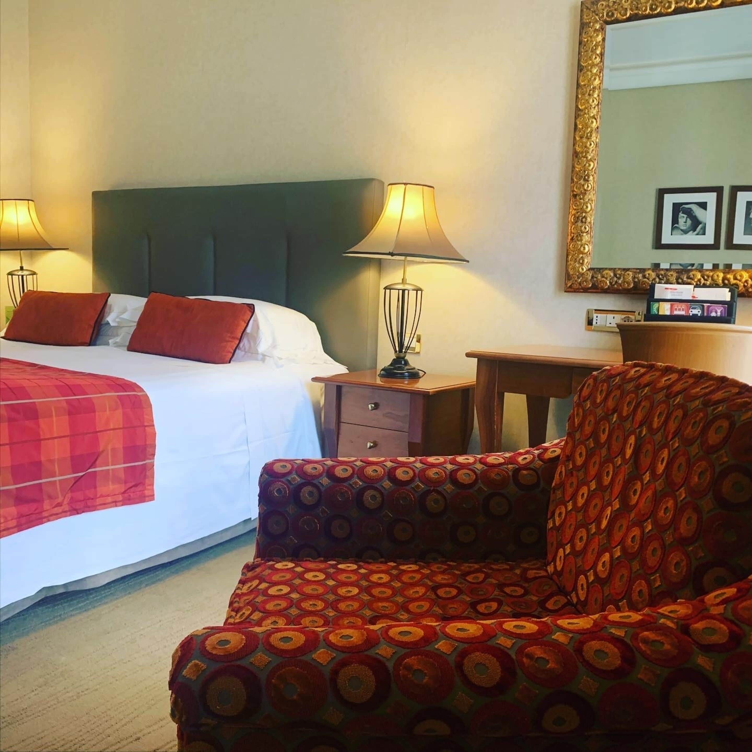 A hotel room with a bed, two bedside lamps, a chair, a nightstand, a dresser, and a mirror on the wall.