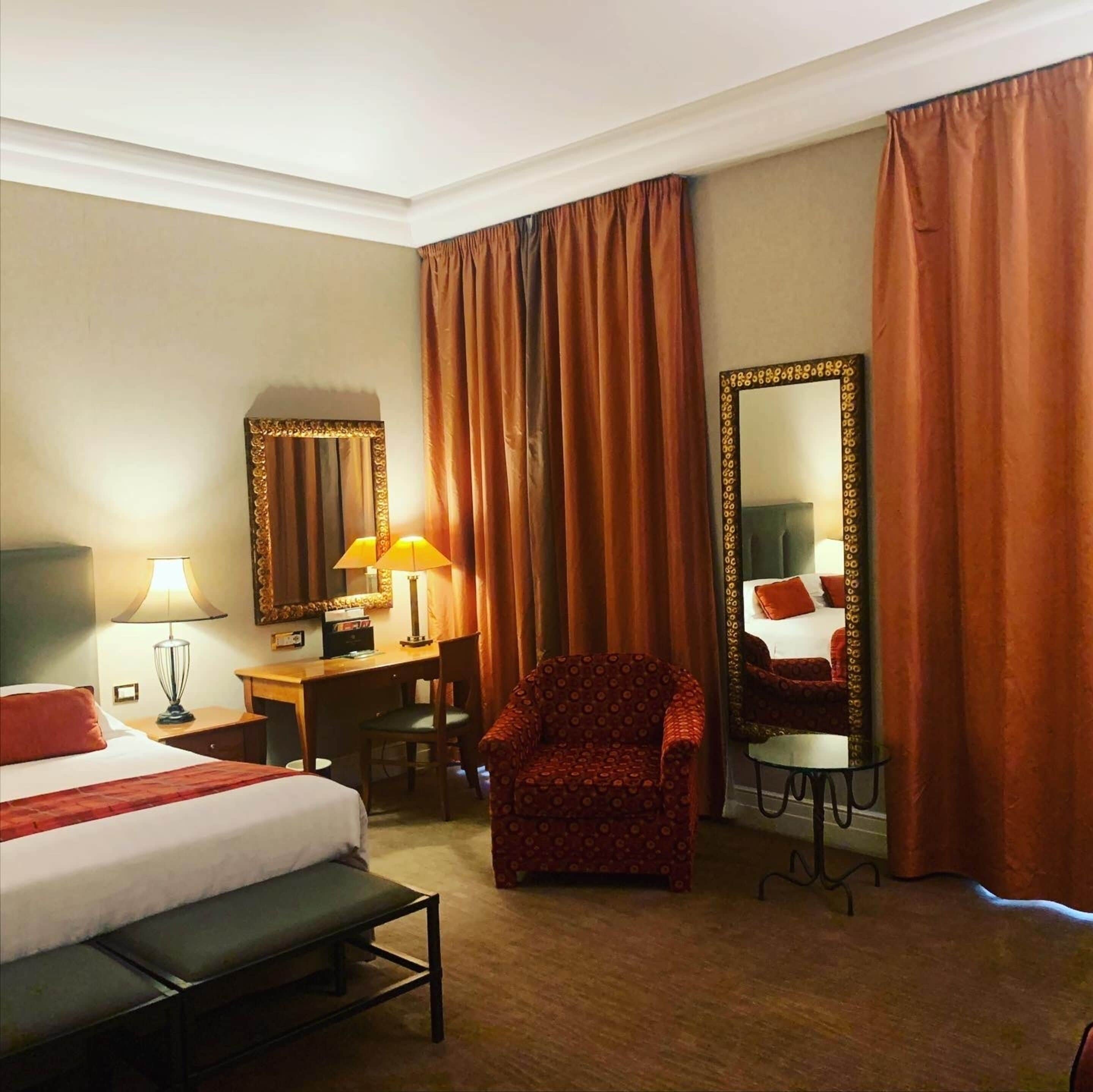 Habitació d'hotel amb llit, escriptori, cadira, làmpades i un mirall gran.