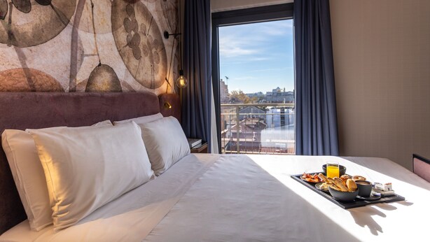 Ein Hotelzimmer mit einem großen Bett, Blick auf die Stadt und Frühstück auf dem Nachttisch.