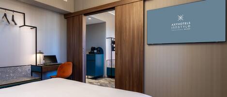 A hotel room with a bed, a desk with a laptop, a chair, and a wardrobe. There is a mirror on the wall and a sign with the hotel name.