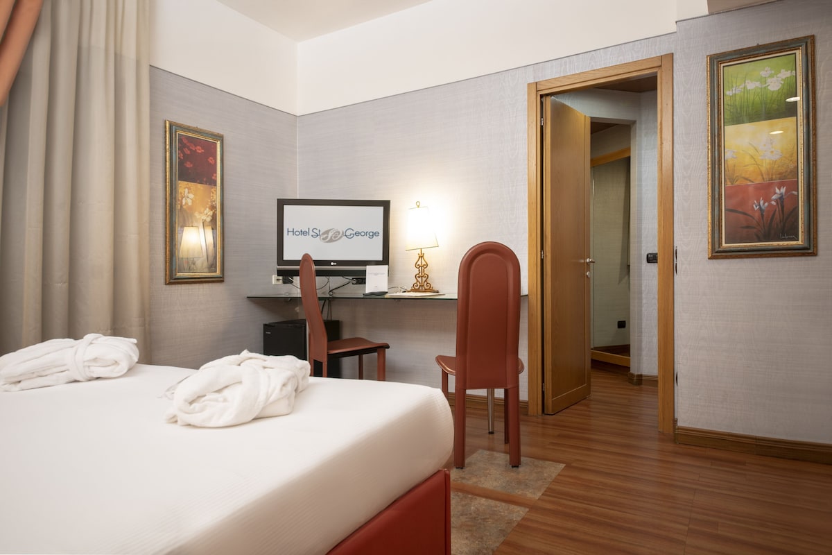A hotel room with a bed, a desk with a computer, a chair, and a television displaying 'Hotel St. George'.