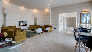 A modern living room with a sofa, chairs, a coffee table, and a TV. There is a dining area with a table and chairs. The room has a light-colored floor and walls with horizontal lines. - Best Western Plus Hotel Farnese (Parma)