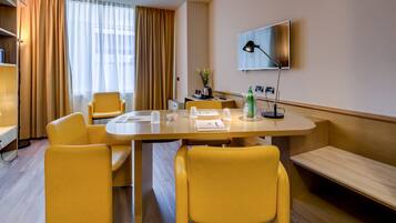 Habitación de hotel con suelo de madera, una mesa de comedor con sillas amarillas, un televisor de pantalla plana y una planta pequeña sobre la mesa.