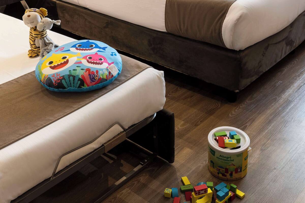 A hotel room with a bed, a bedside table, a plush toy, a colorful pillow, and scattered building blocks on the floor.
