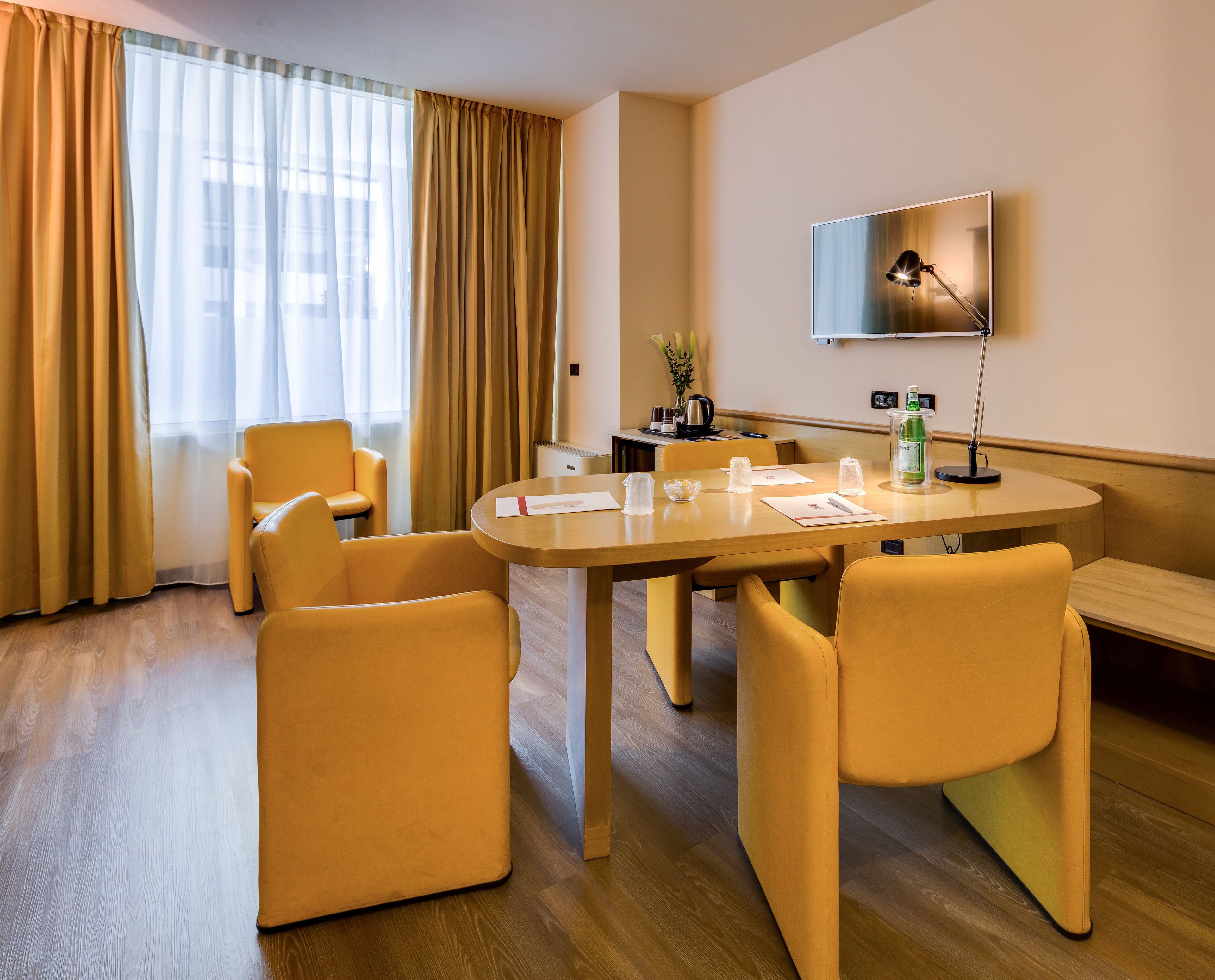 A hotel room with a round table, four yellow chairs, a flat-screen TV mounted on the wall, and a wooden floor.