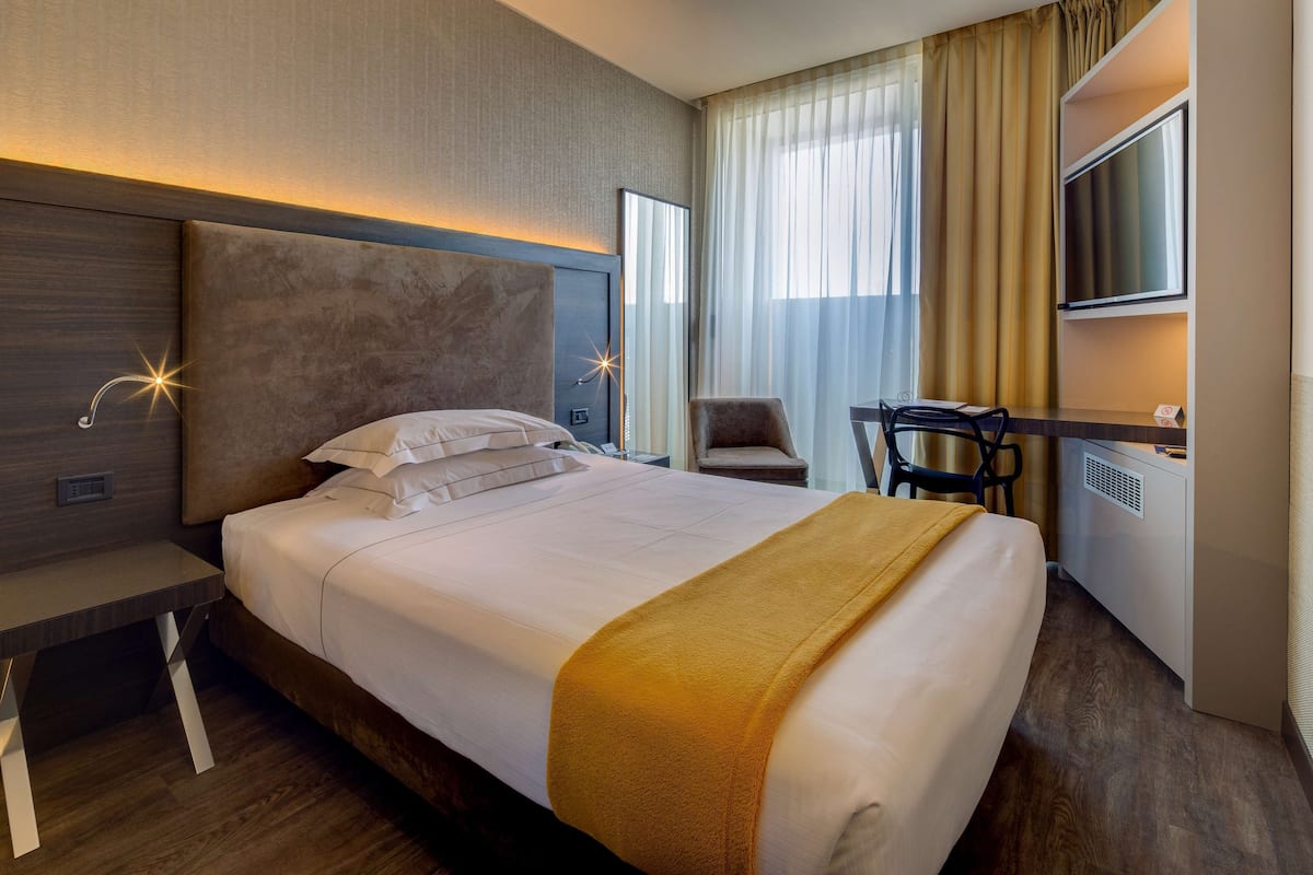 A hotel room with a large bed, a desk, a chair, a TV, and a window with curtains.