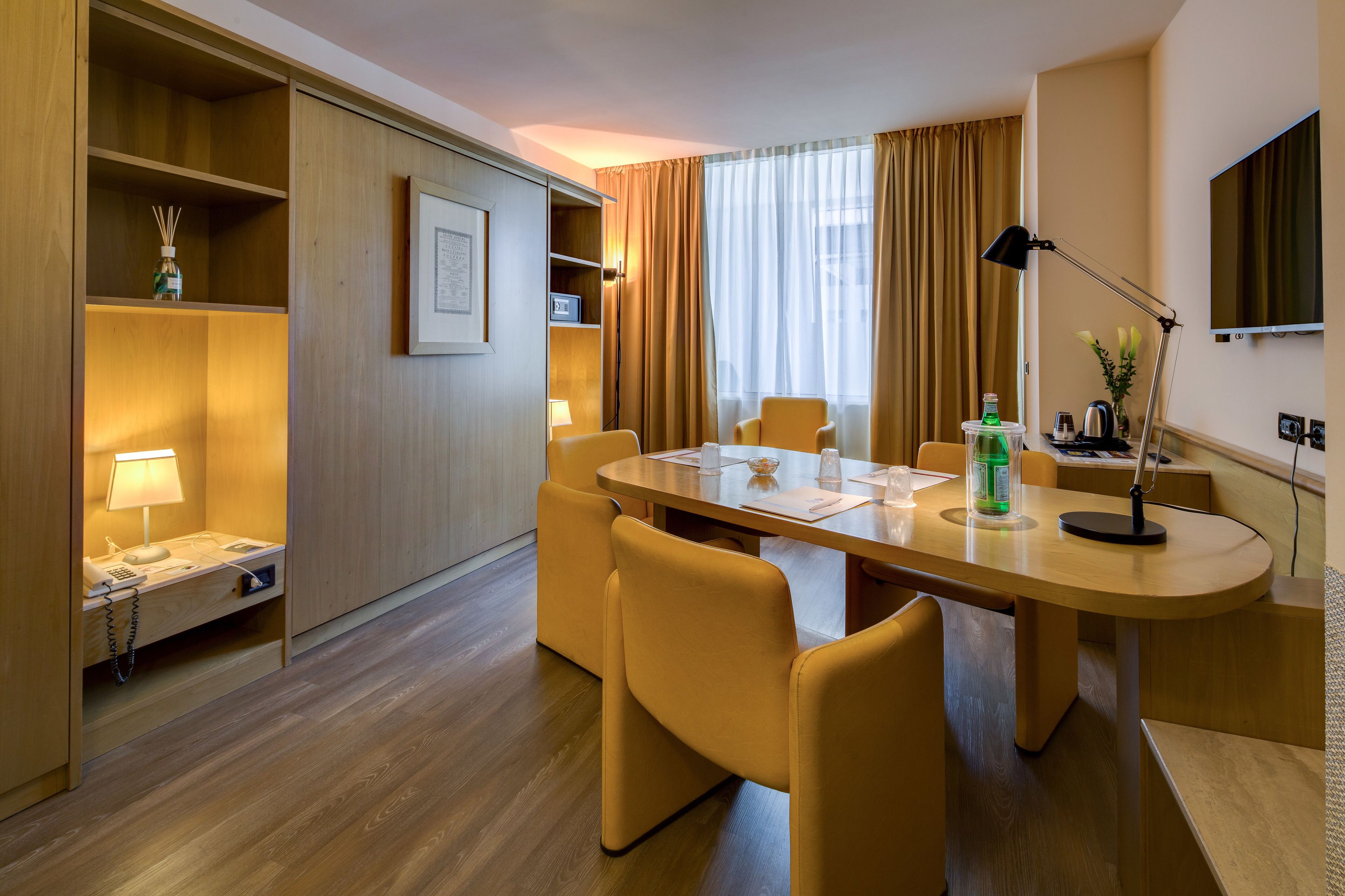 A hotel room with a wooden desk, a telephone, a lamp, a chair, a table, a television, and a window with curtains.