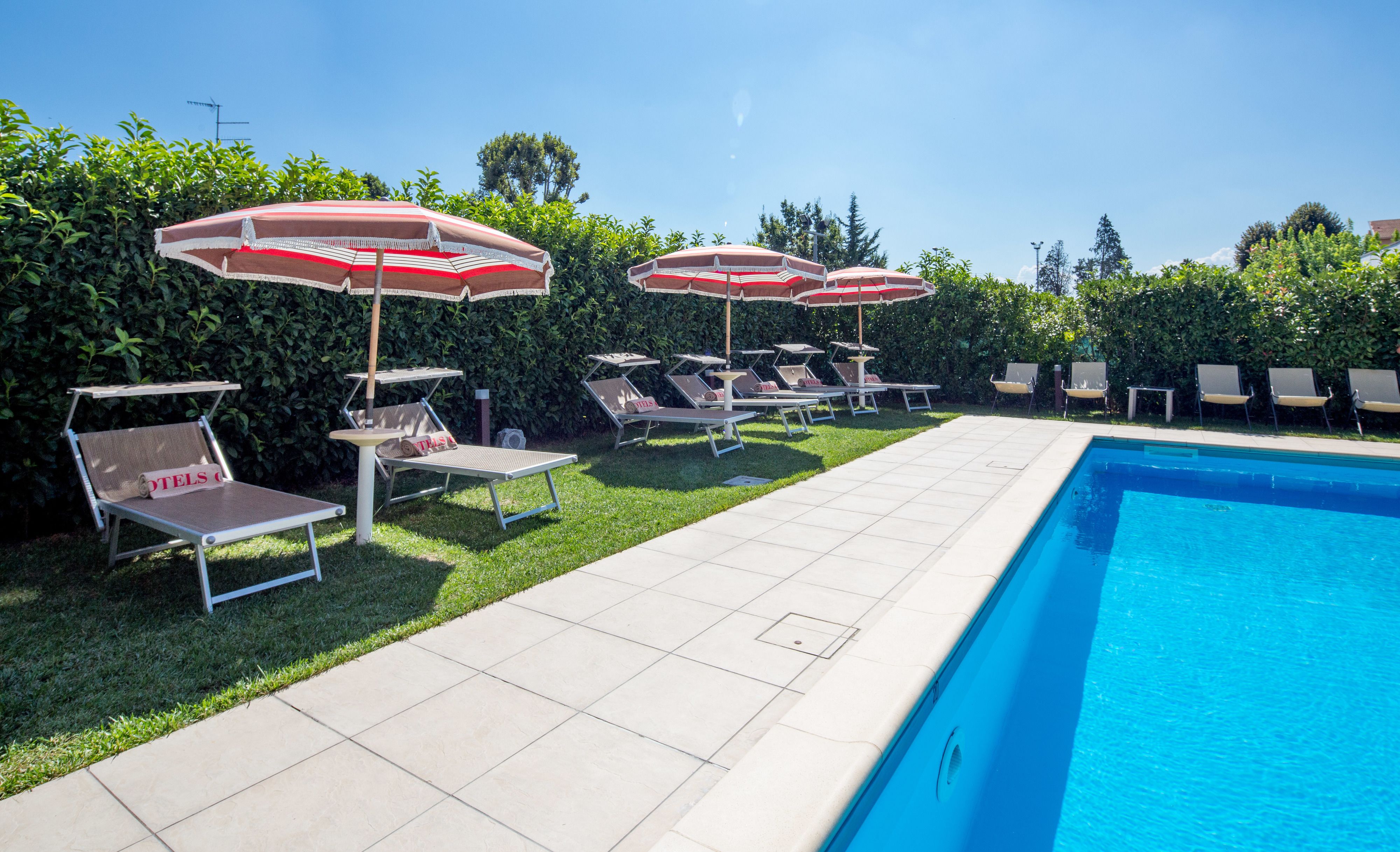 Uma área à beira da piscina com espreguiçadeiras e guarda-sóis, uma piscina azul cristalina e um gramado bem cuidado.