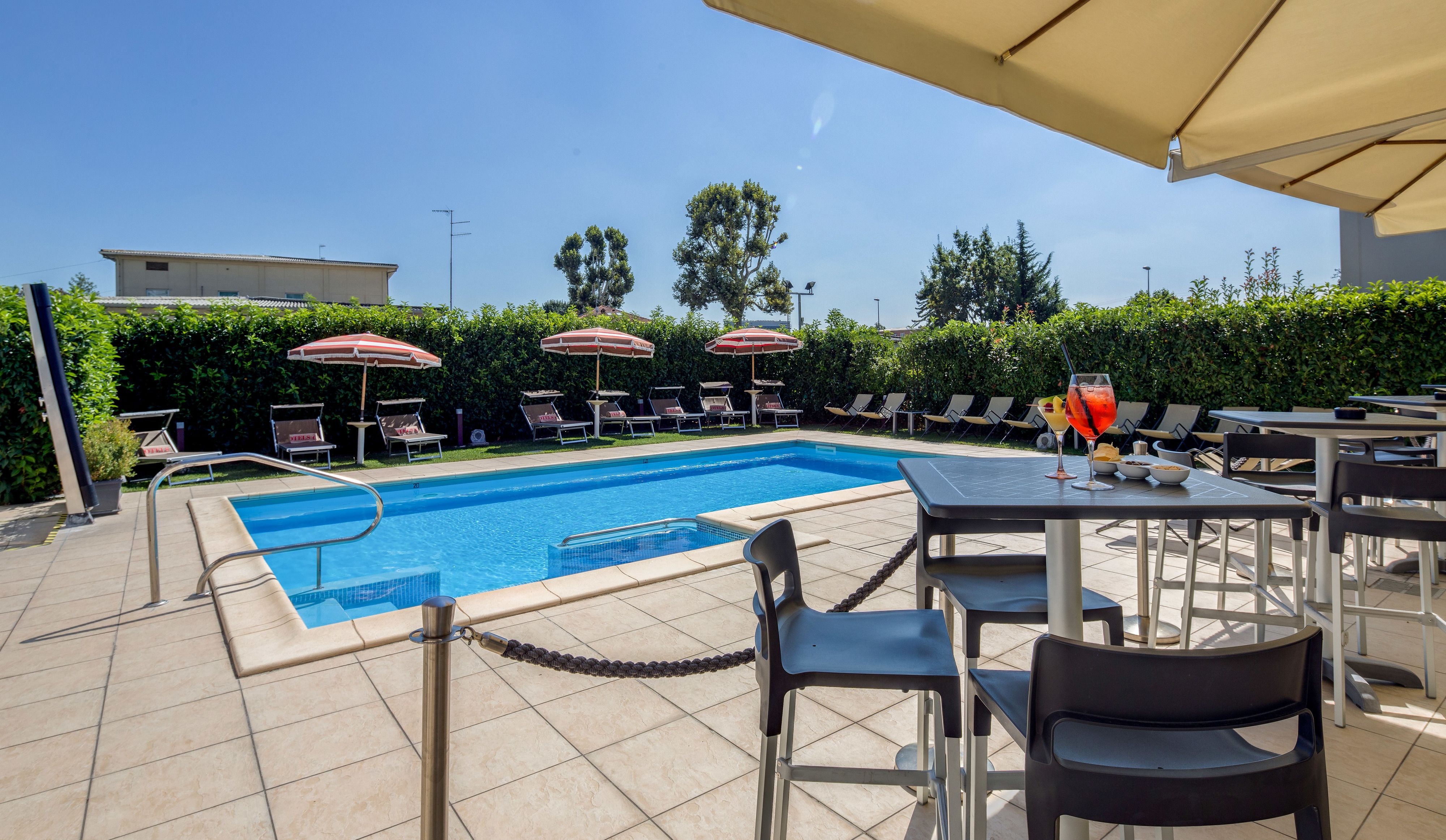 Un espace piscine avec des chaises longues et des parasols, une table à manger avec des chaises, et un ciel d’un bleu éclatant.