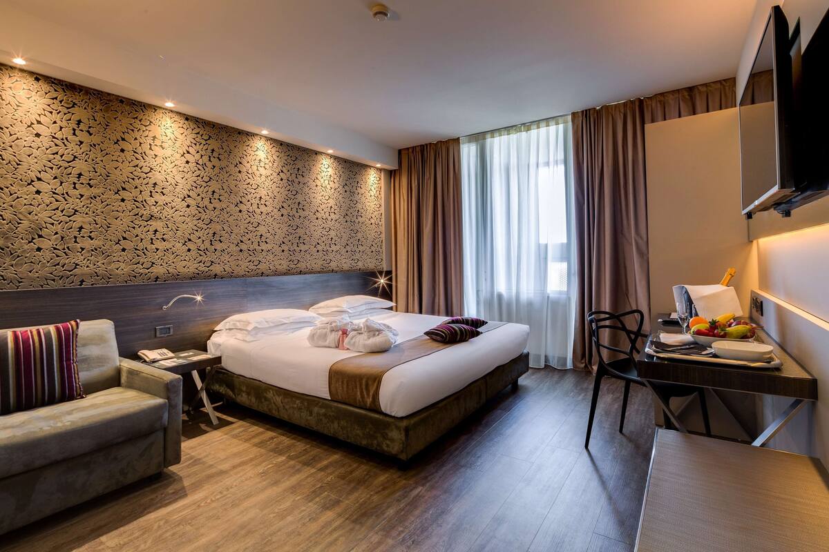 A hotel room with a large bed, a desk with a chair, a sofa, and a TV mounted on the wall.