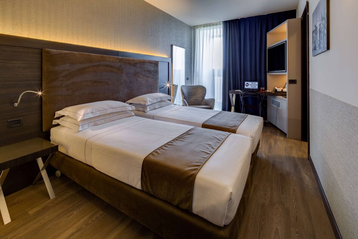 A hotel room with two beds, a desk, a chair, a TV, and a large window with curtains.