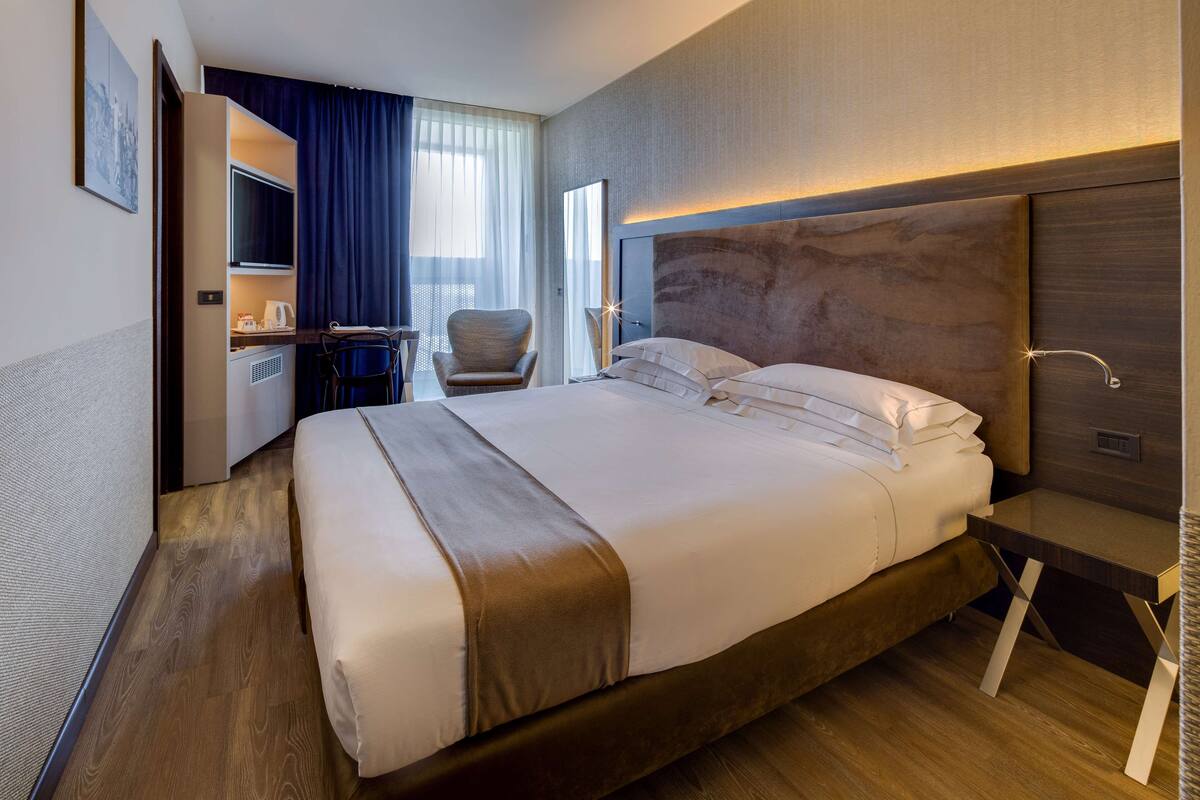 A hotel room with a large bed, a desk with a chair, a television, and a window with curtains.