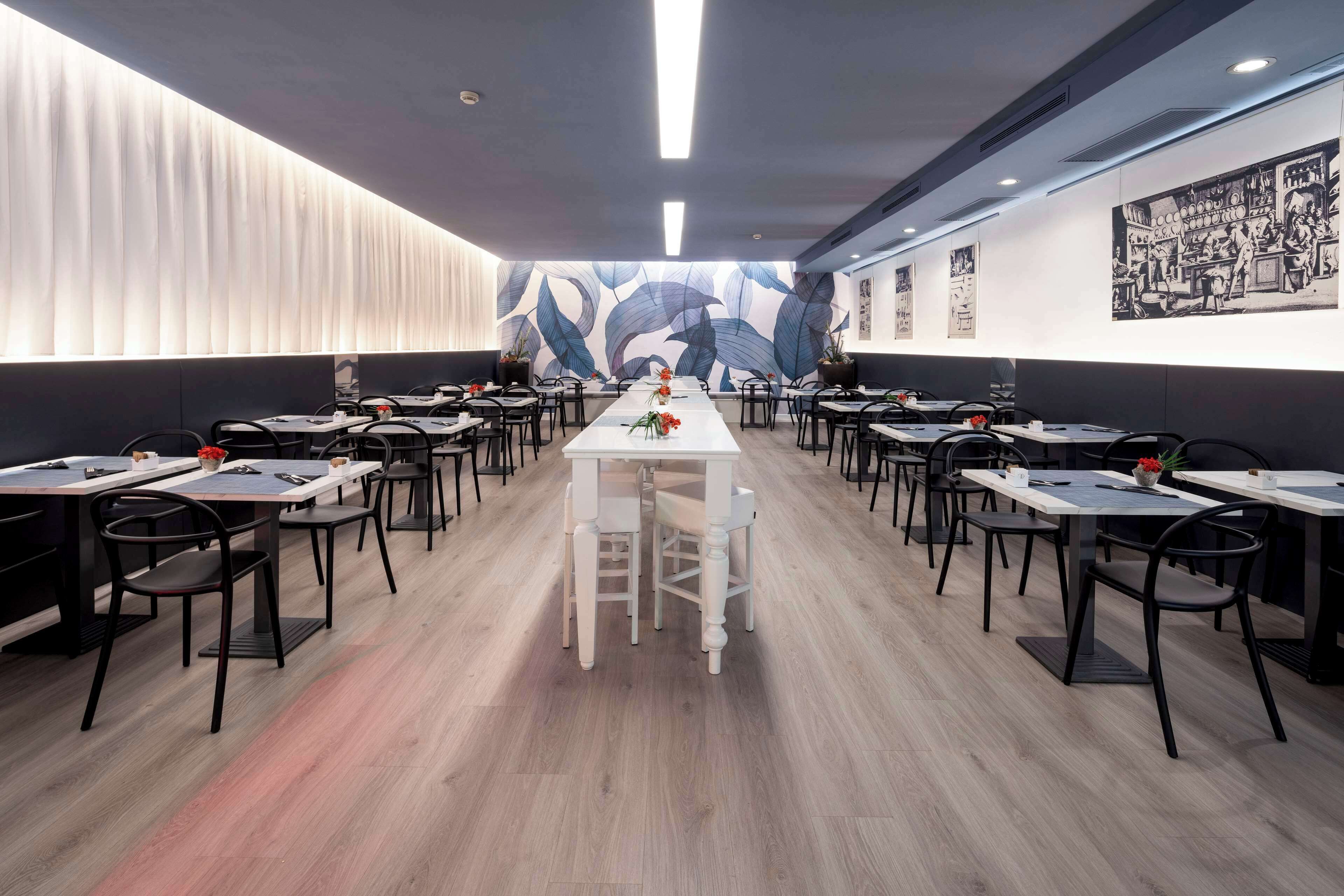 A modern dining area with long tables and chairs, a central white table with a vase of flowers, and a wall mural with abstract leaf patterns.