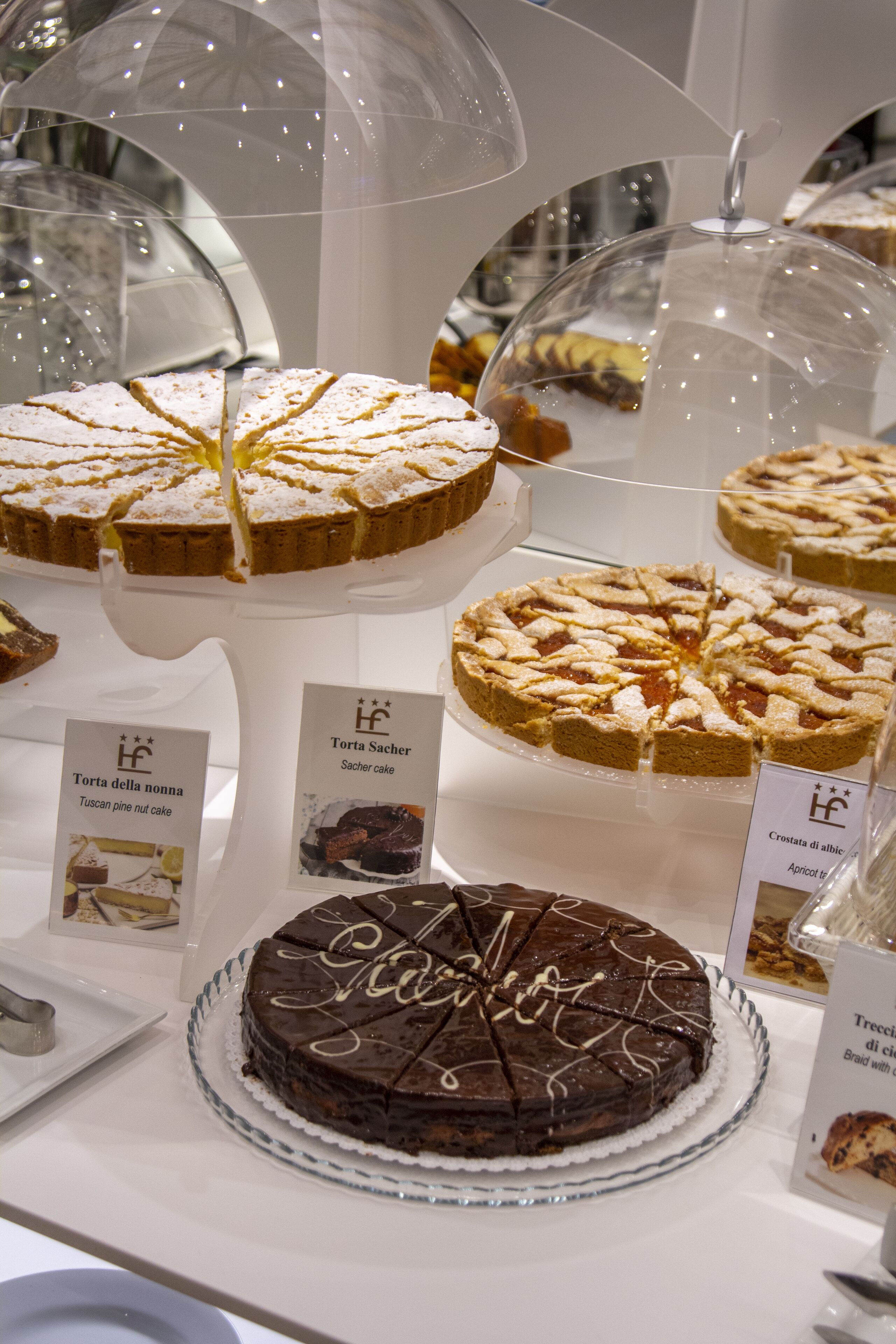 A display of various cakes under glass domes, including a chocolate cake with a lattice design.