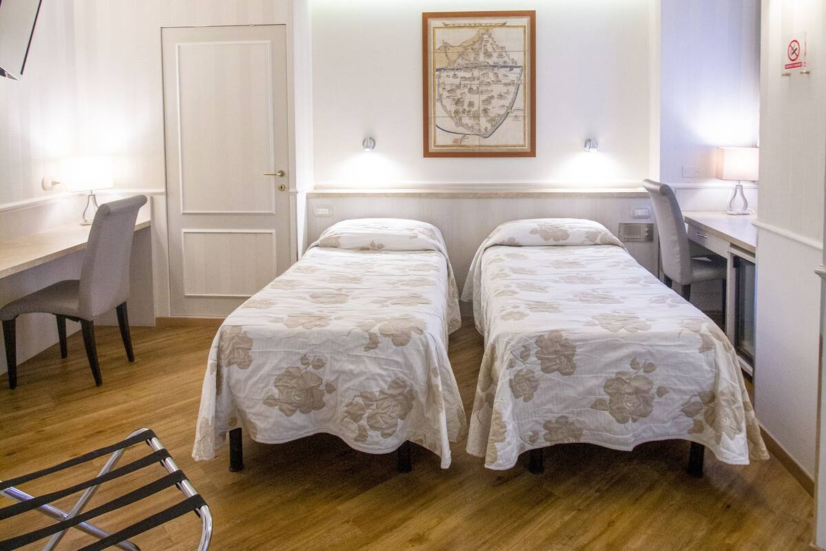 A hotel room with two single beds, a desk, a chair, a lamp, and a map on the wall.