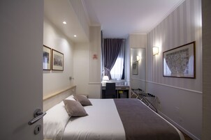 A hotel room with a bed, a desk, a chair, a lamp, a map on the wall, and a door.