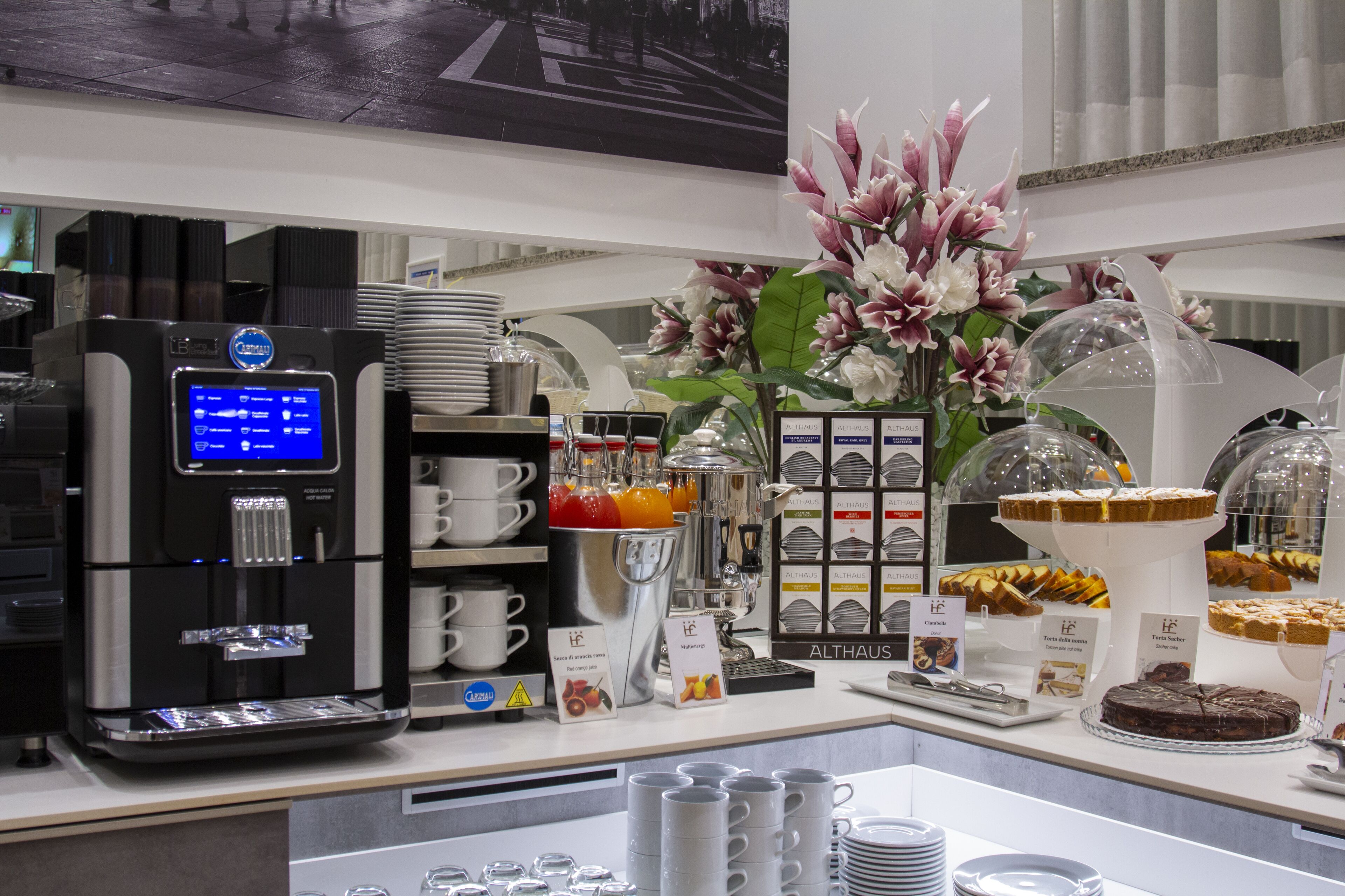 Zona de desayuno tipo bufé con una máquina de café, diferentes opciones de comida y un centro de mesa floral.