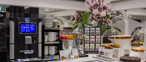 Zona de desayuno tipo bufé con una máquina de café, diferentes opciones de comida y un centro de mesa floral.
