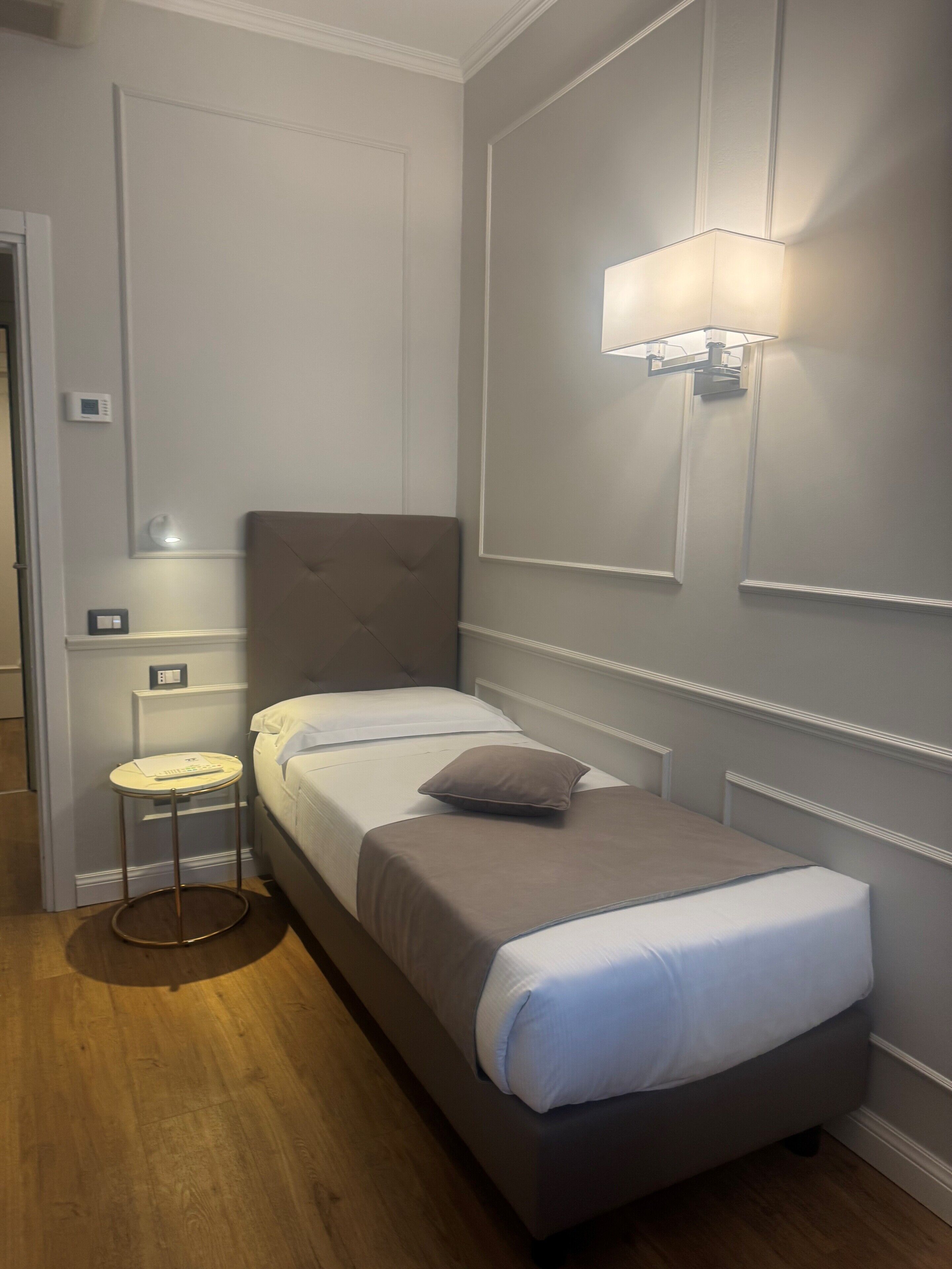A single bed with a headboard, a bedside table with a lamp, and a wall-mounted light fixture in a room with wooden flooring.