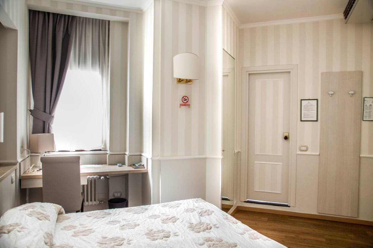 A hotel room with a bed, a desk, a chair, a window with curtains, a wall-mounted lamp, a door, and a radiator.