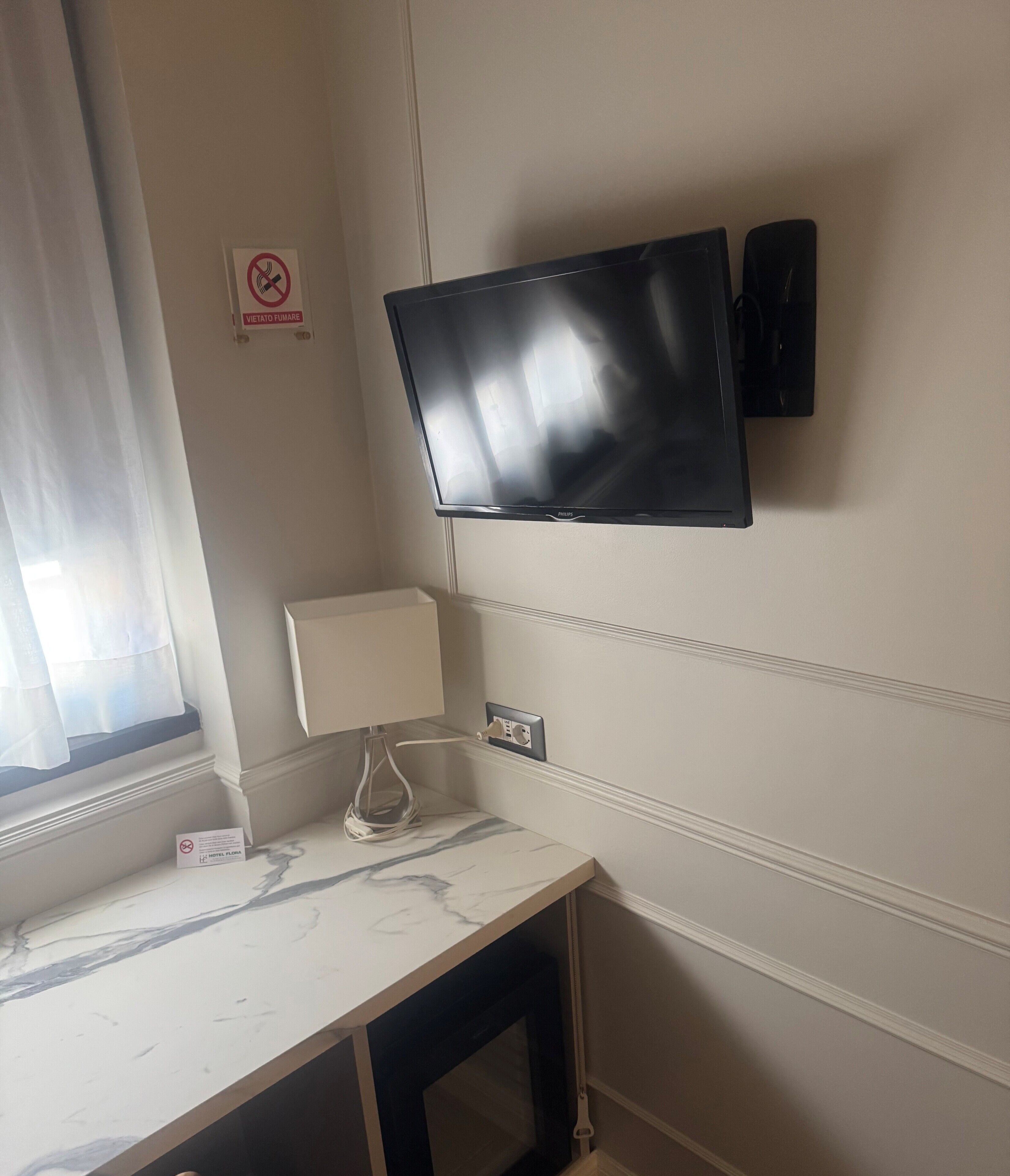 A hotel room with a flat-screen TV mounted on the wall, a marble-patterned desk, a lamp, and a no-smoking sign.