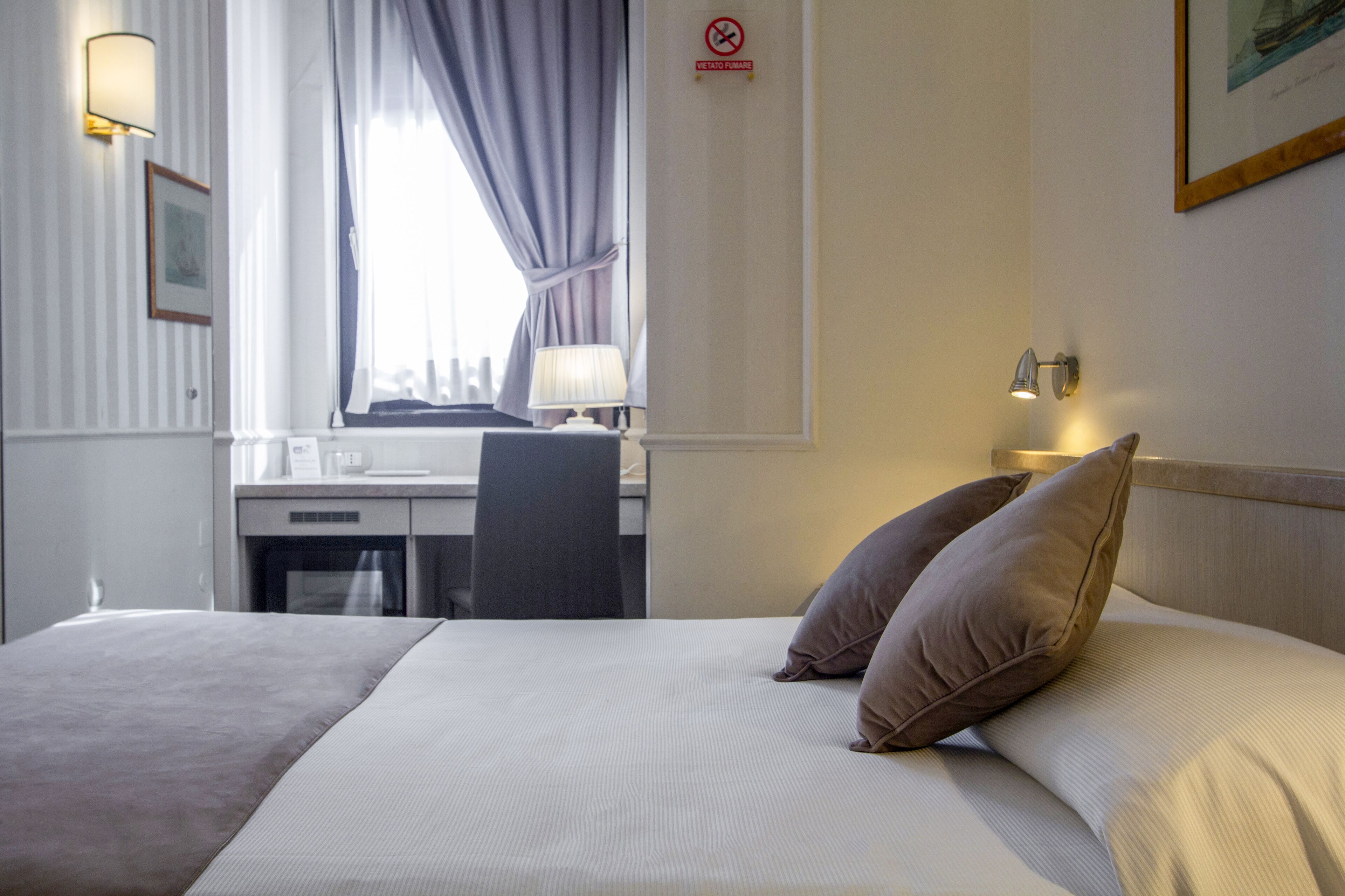 Ein Hotelzimmer mit einem Bett, einem Schreibtisch und einem Stuhl. Es gibt ein Fenster mit Vorhängen, eine Lampe auf dem Schreibtisch und ein Schild an der Wand.