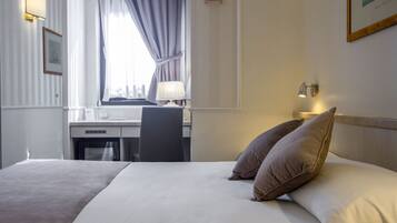 Ein Hotelzimmer mit einem Bett, einem Schreibtisch und einem Stuhl. Es gibt ein Fenster mit Vorhängen, eine Lampe auf dem Schreibtisch und ein Schild an der Wand.