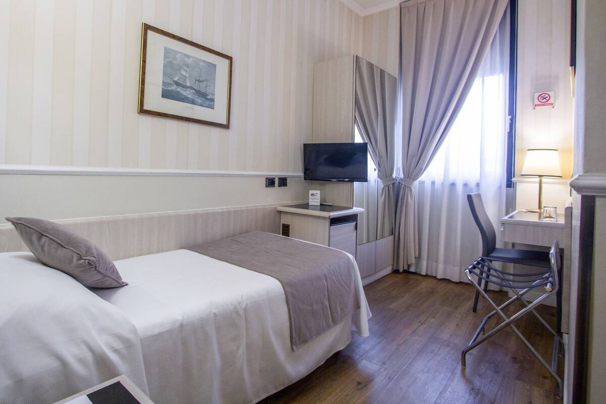 A hotel room with a bed, a desk with a television, a chair, and a window with curtains.