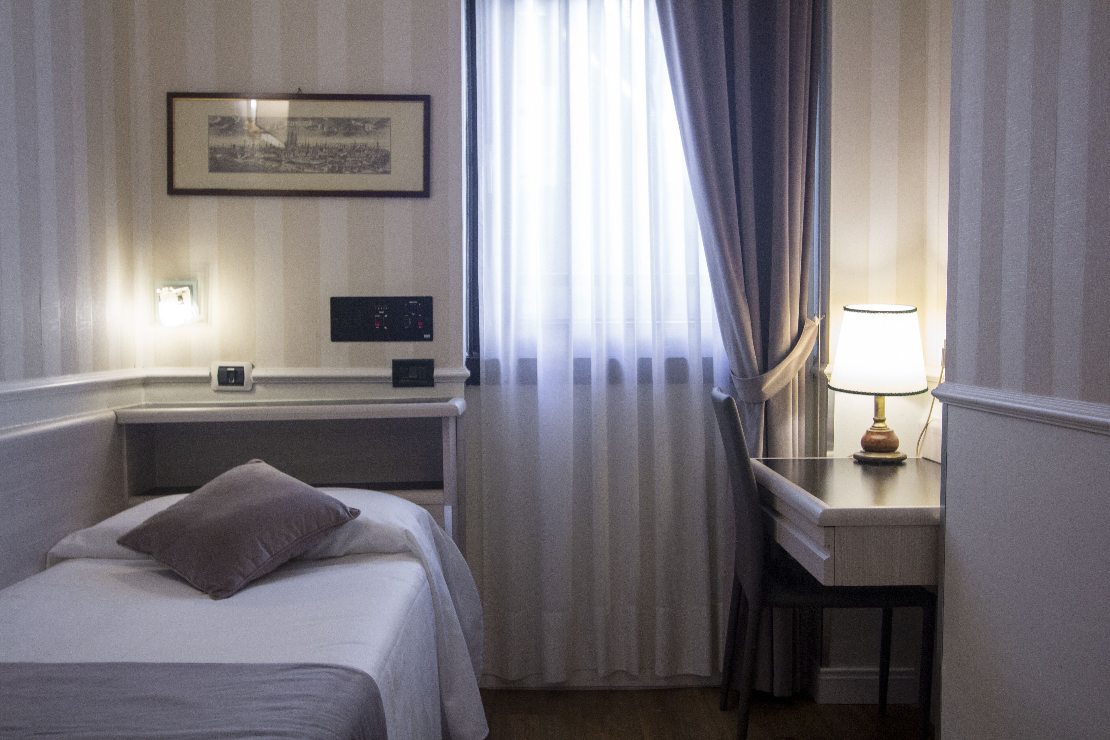A hotel room with a bed, a desk, a lamp, a picture on the wall, and a window with curtains.