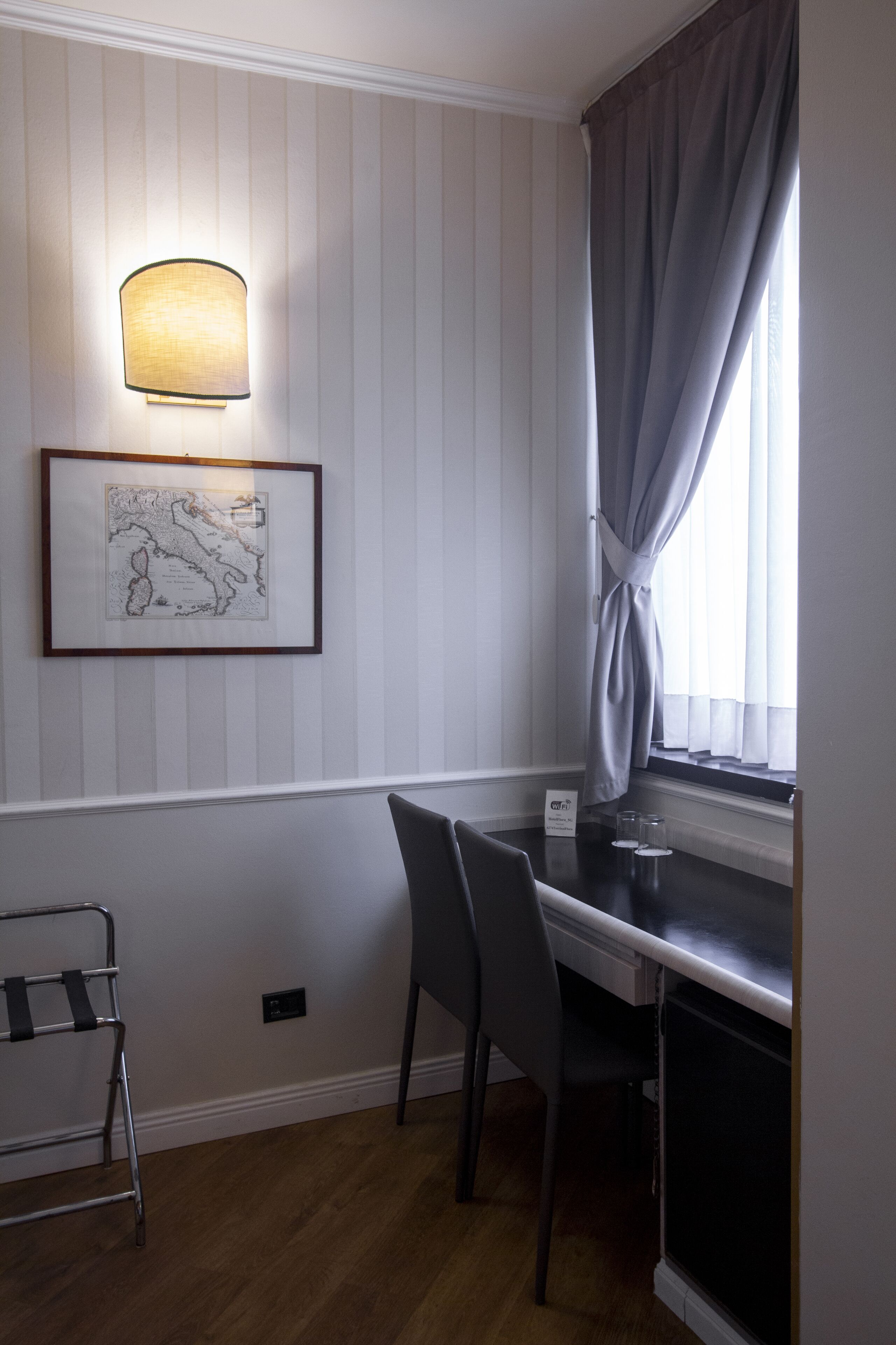A room with a dining table, chairs, a lamp, a framed map, and a window with curtains.
