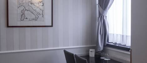 A room with a dining table, chairs, a lamp, a framed map, and a window with curtains.